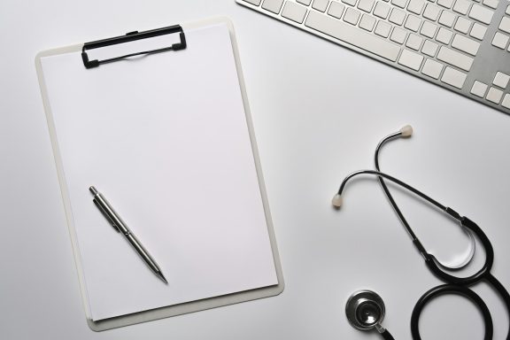A blank clipboard with a pen and a stethoscope beside a keyboard.